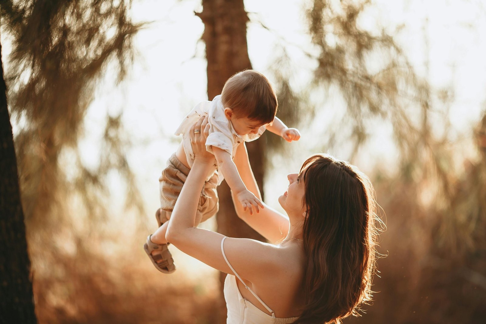 A mother joyfully lifts her smiling child outdoors in the warm, golden sunlight, surrounded by trees.